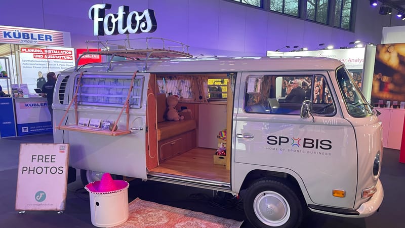 Ein cremeweißer Fotobulli steht als eigenständiger Messestand: geöffnete Schiebetür mit Sitzbank, davor ein Aufsteller mit „Free Photos“ und ein Teppich.