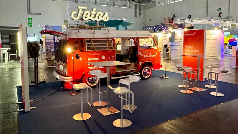 Ein rot folierter Fotobulli steht als Blickfang mitten im Messestand der Agentur BRANDUNG: blauer Standteppich, Stehtisch mit Barhockern und eine Markenwand rechts.