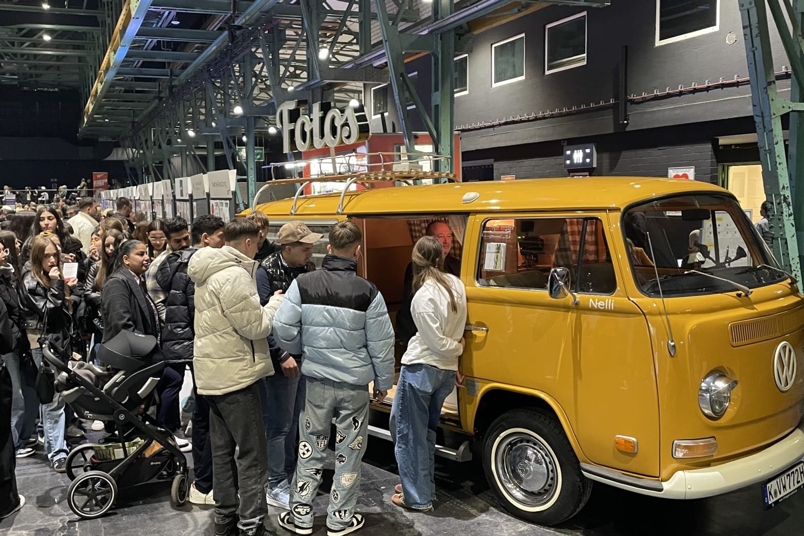 Karrieretag München: Eine Schlange junger Kandidatinnen und Kandidaten wartet in der Messehalle vor dem orangefarbenen Fotobulli mit beleuchtetem „Fotos“-Schriftzug auf dem Dach.