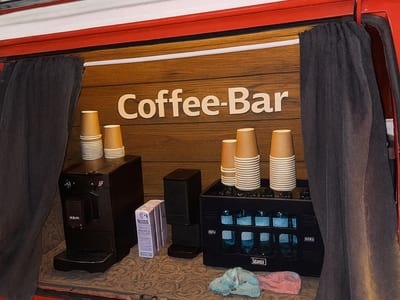 Eine geöffnete Klappe des Bullis mit Holzschild „Coffee-Bar“, Espressomaschine und gestapelten Bechern.